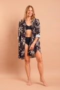A modelo Carla Barros sorri em uma fotografia de corpo inteiro, vestindo um conjunto da marca Isa Rodrigues Resort Wear. Ela usa um biquíni preto texturizado de cintura alta e um top combinando, sob uma camisa de botão longa e fluida com estampa botânica branca e cinza.  Ela está descalça contra um fundo bege liso e uniforme.