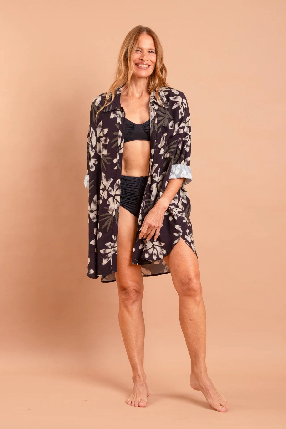 A modelo Carla Barros sorri em uma fotografia de corpo inteiro, vestindo um conjunto da marca Isa Rodrigues Resort Wear. Ela usa um biquíni preto texturizado de cintura alta e um top combinando, sob uma camisa de botão longa e fluida com estampa botânica branca e cinza.  Ela está descalça contra um fundo bege liso e uniforme.