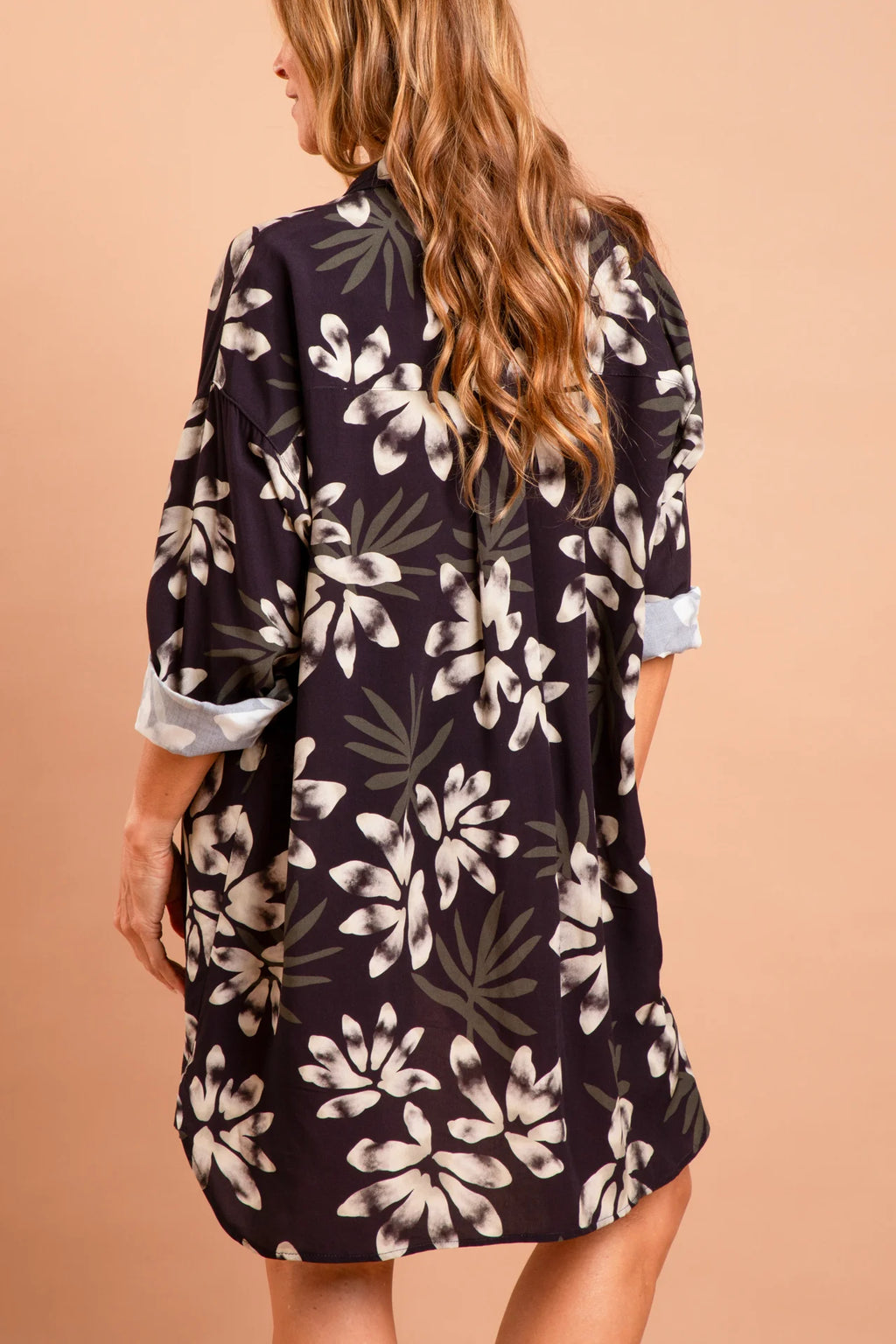 A modelo Carla Barros de costas vestindo um conjunto da marca Isa Rodrigues Resort Wear. Ela usa uma camisa longa e fluida com estampa botânica branca e cinza.  Ela está contra um fundo bege liso e uniforme.