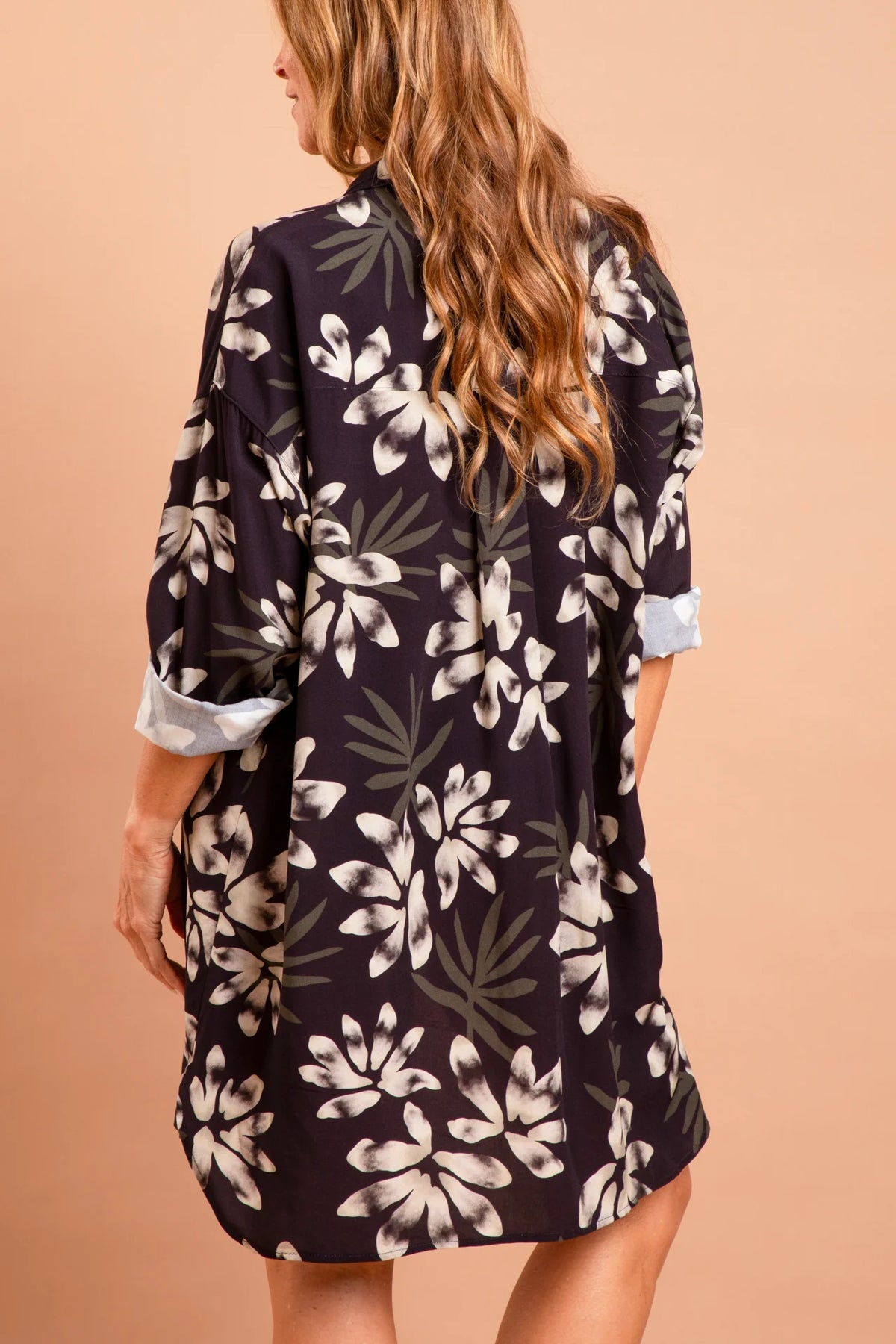 A modelo Carla Barros de costas vestindo um conjunto da marca Isa Rodrigues Resort Wear. Ela usa uma camisa longa e fluida com estampa botânica branca e cinza.  Ela está contra um fundo bege liso e uniforme.