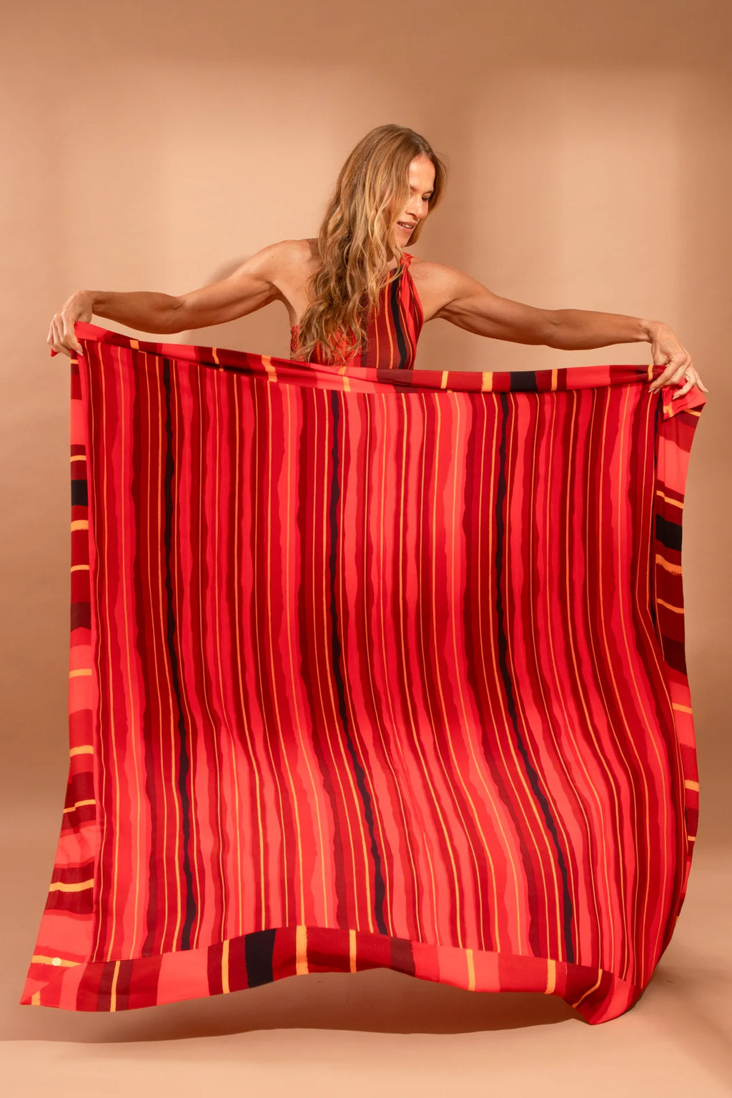 Fotografia de canga de praia quadrada ampla em viscose com padronagem geométrica de listras estreitas e barrado largo, marca Isa Rodrigues Resortwear. A Modelo mostra a canga